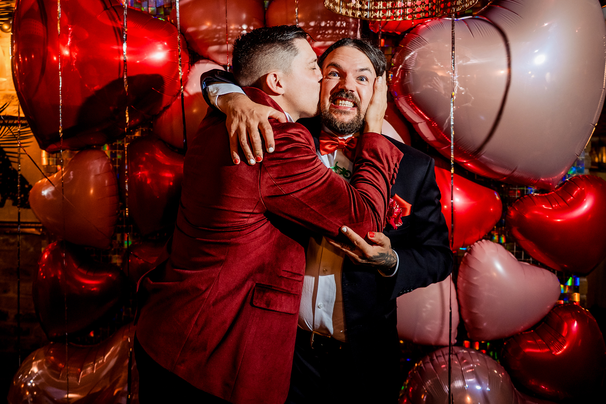 Guest kisses the groom's cheek in front of the heart balloon backdrop.