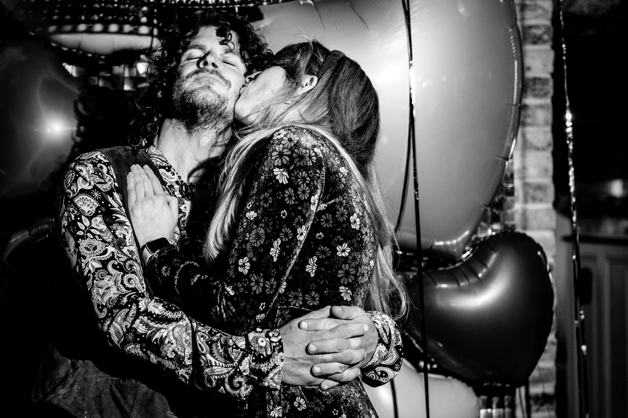Guest kisses her partner's cheek while they hug in front of heart balloons.