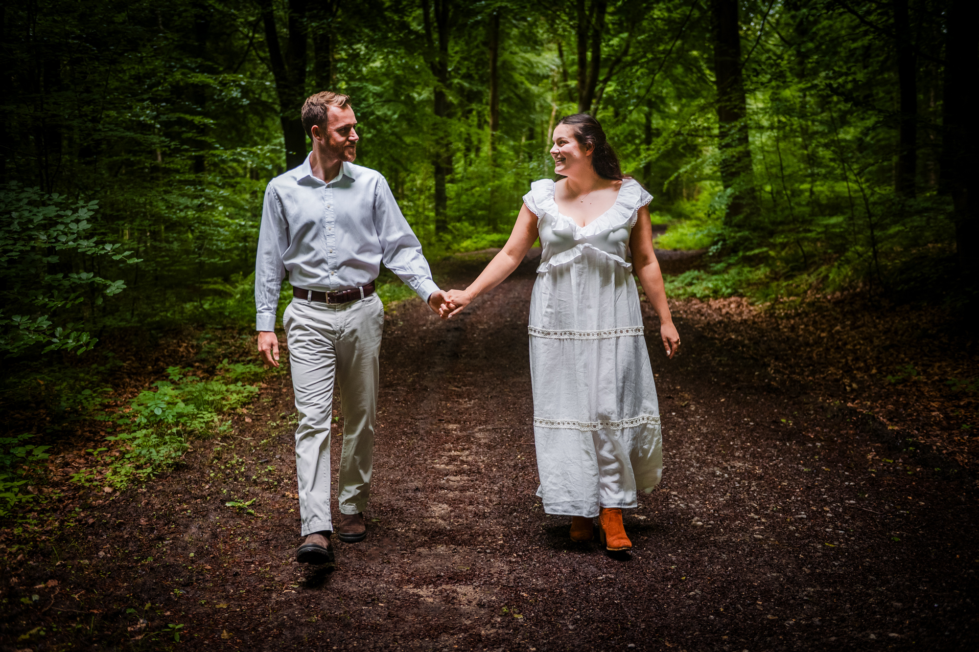 Couple holding hands and walking along a forest path during a West Woods engagement session.