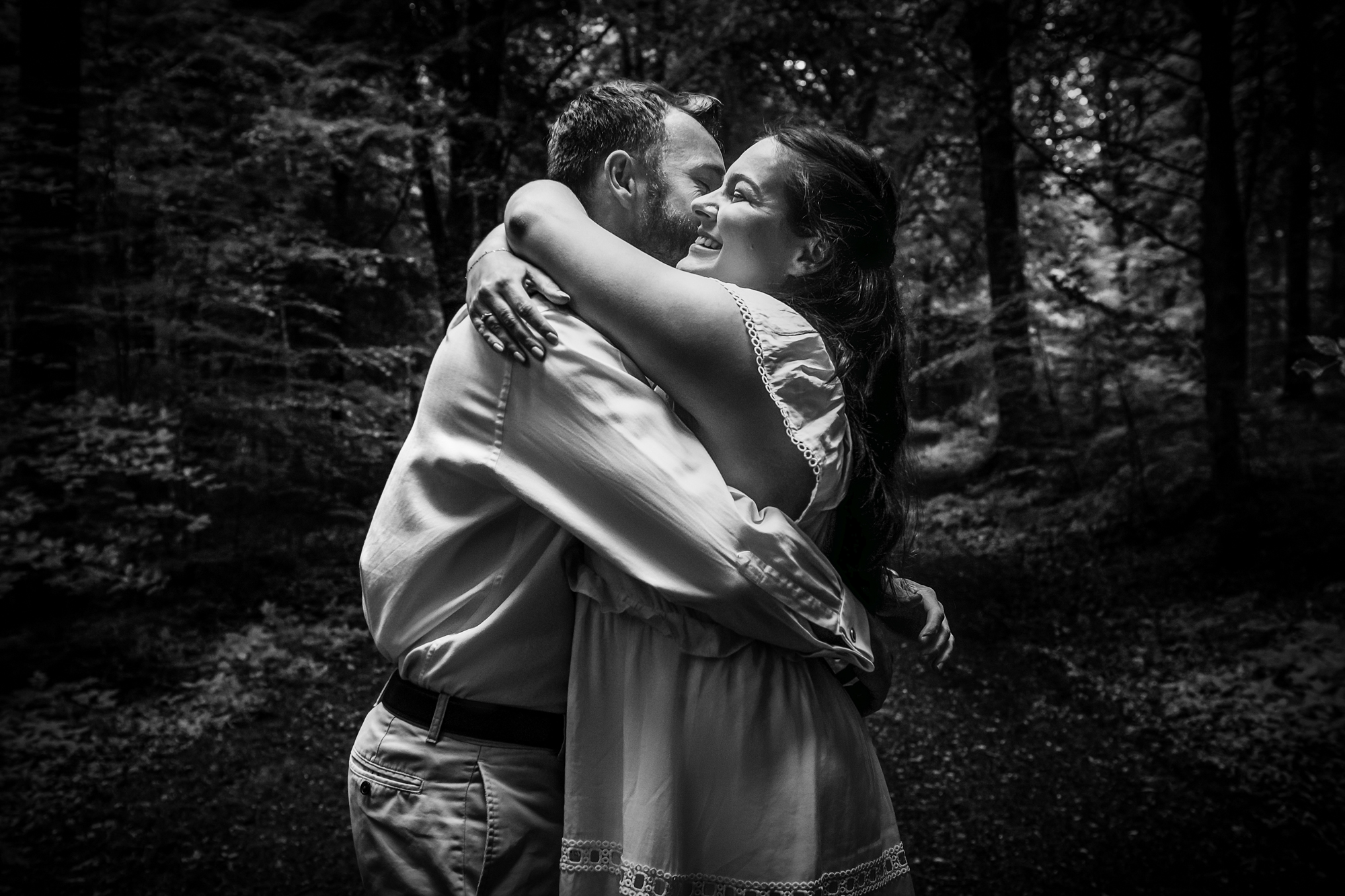 Couple hugging and smiling in a moody black and white portrait among tall trees.