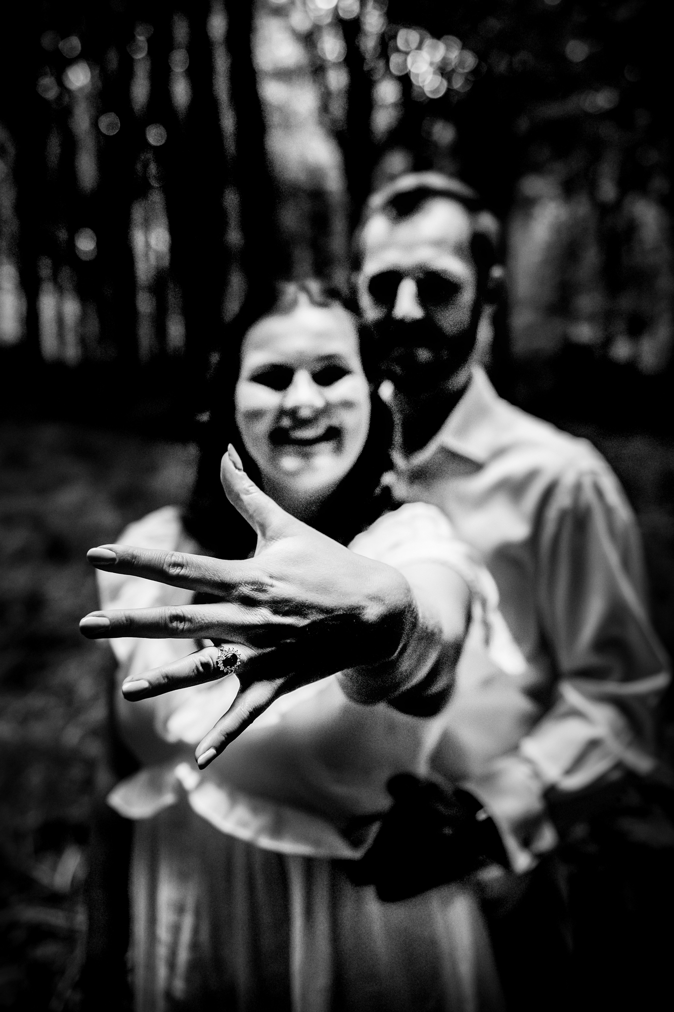 Black and white close-up of an engagement ring held toward the camera in the forest.