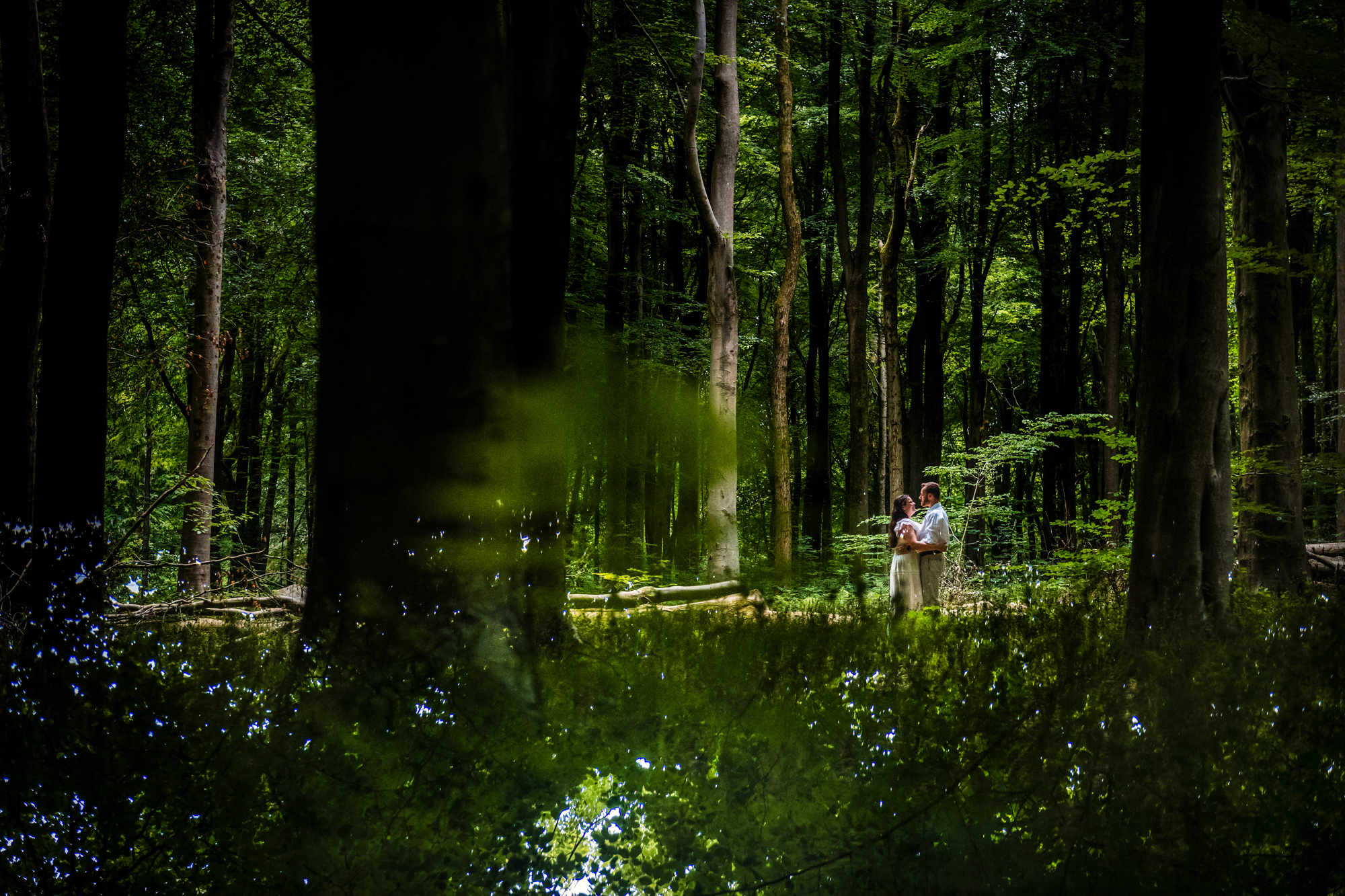 Couple standing together in a forest clearing, surrounded by deep green trees.