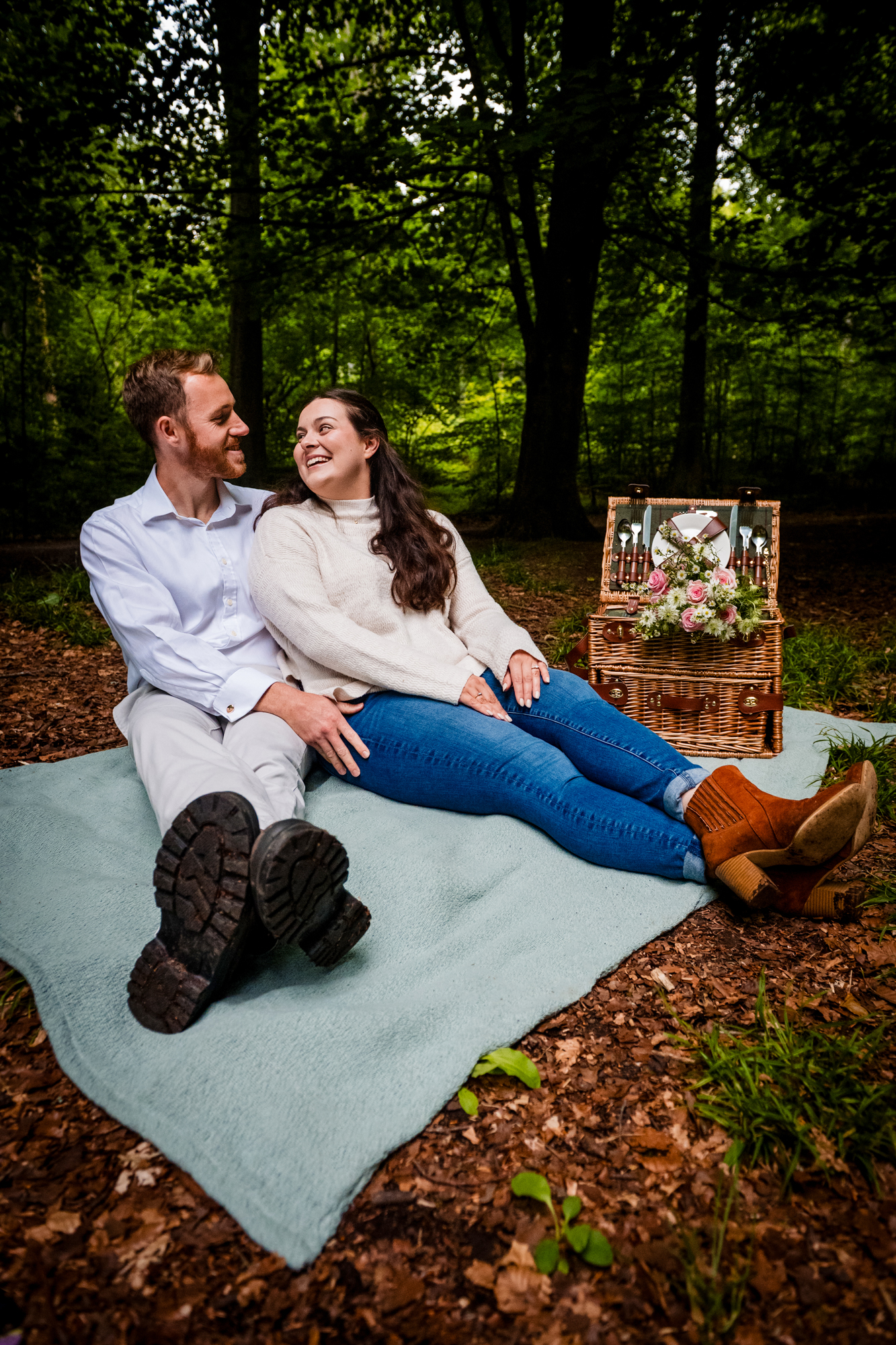 Couple sitting on a blanket beside a picnic setup during their West Woods engagement session.