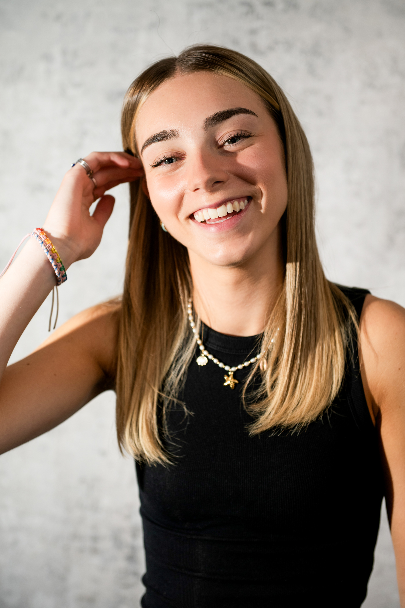 Relaxed studio portrait of smiling woman in black sleeveless top against textured grey backdrop.