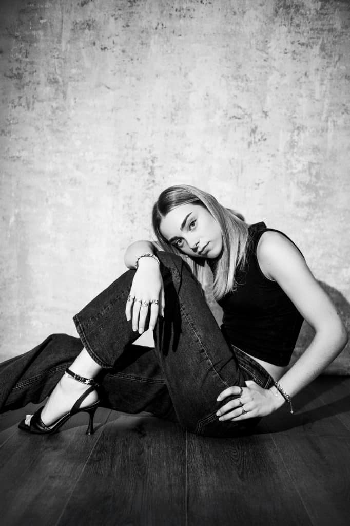 Black and white fashion portrait of seated young woman wearing black heels and dark denim jeans.