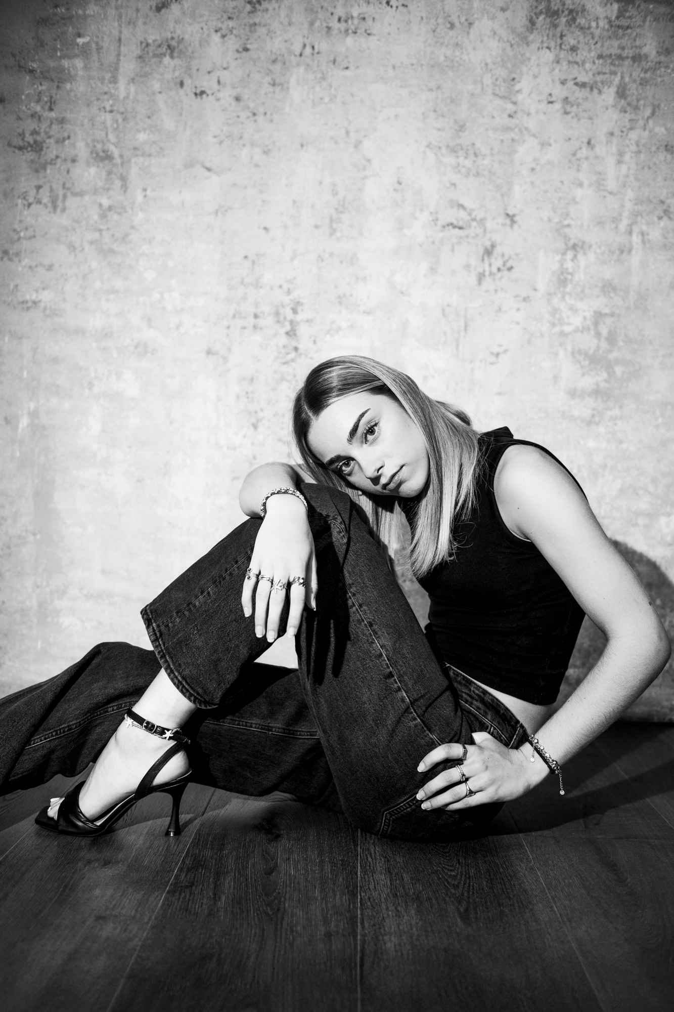 Black and white fashion portrait of seated young woman wearing black heels and dark denim jeans.