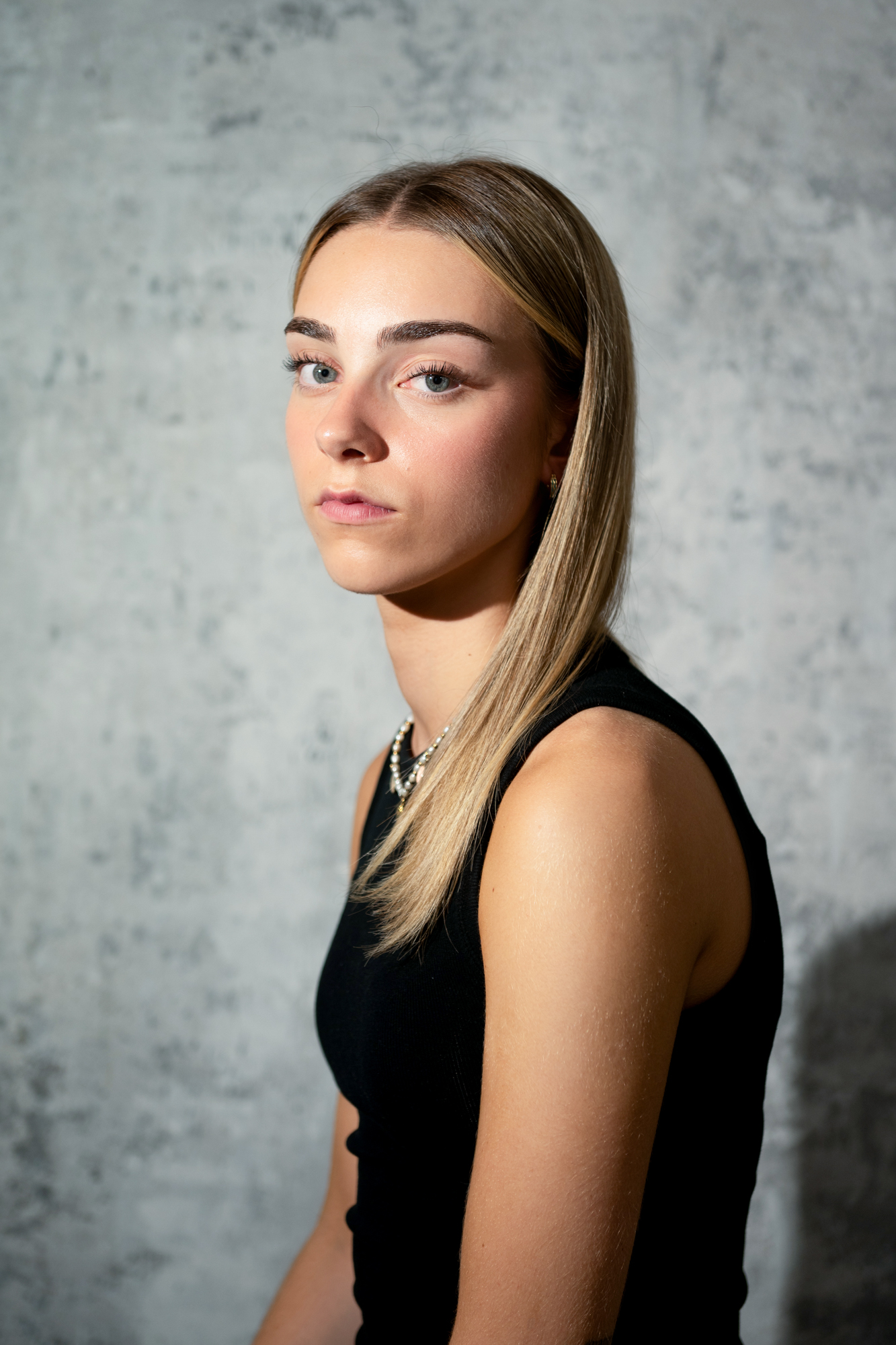 Softly lit side portrait of young woman in black sleeveless top against textured grey studio backdrop.
