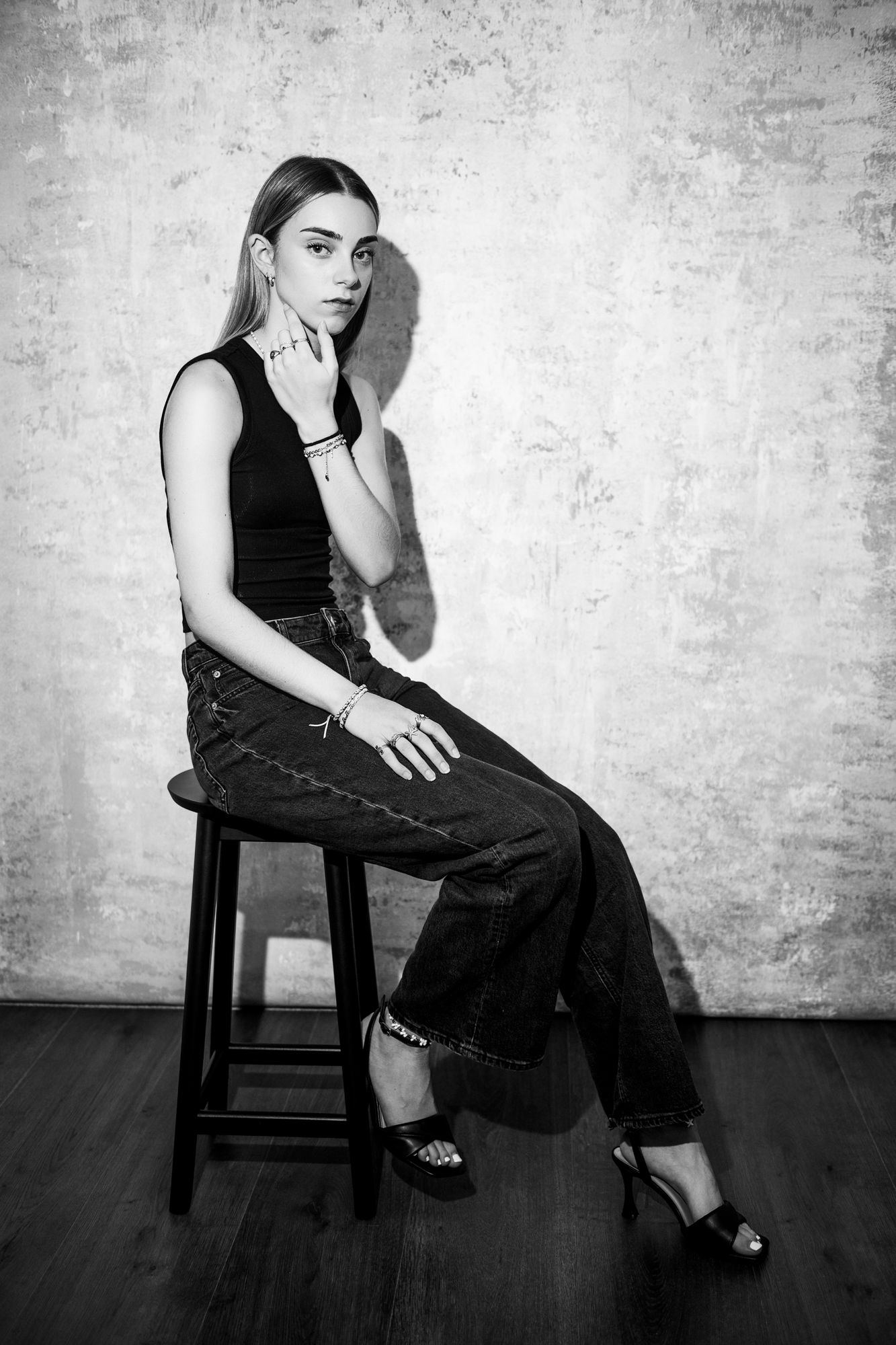 Black and white studio portrait of seated young woman with dramatic shadow and textured wall backdrop.