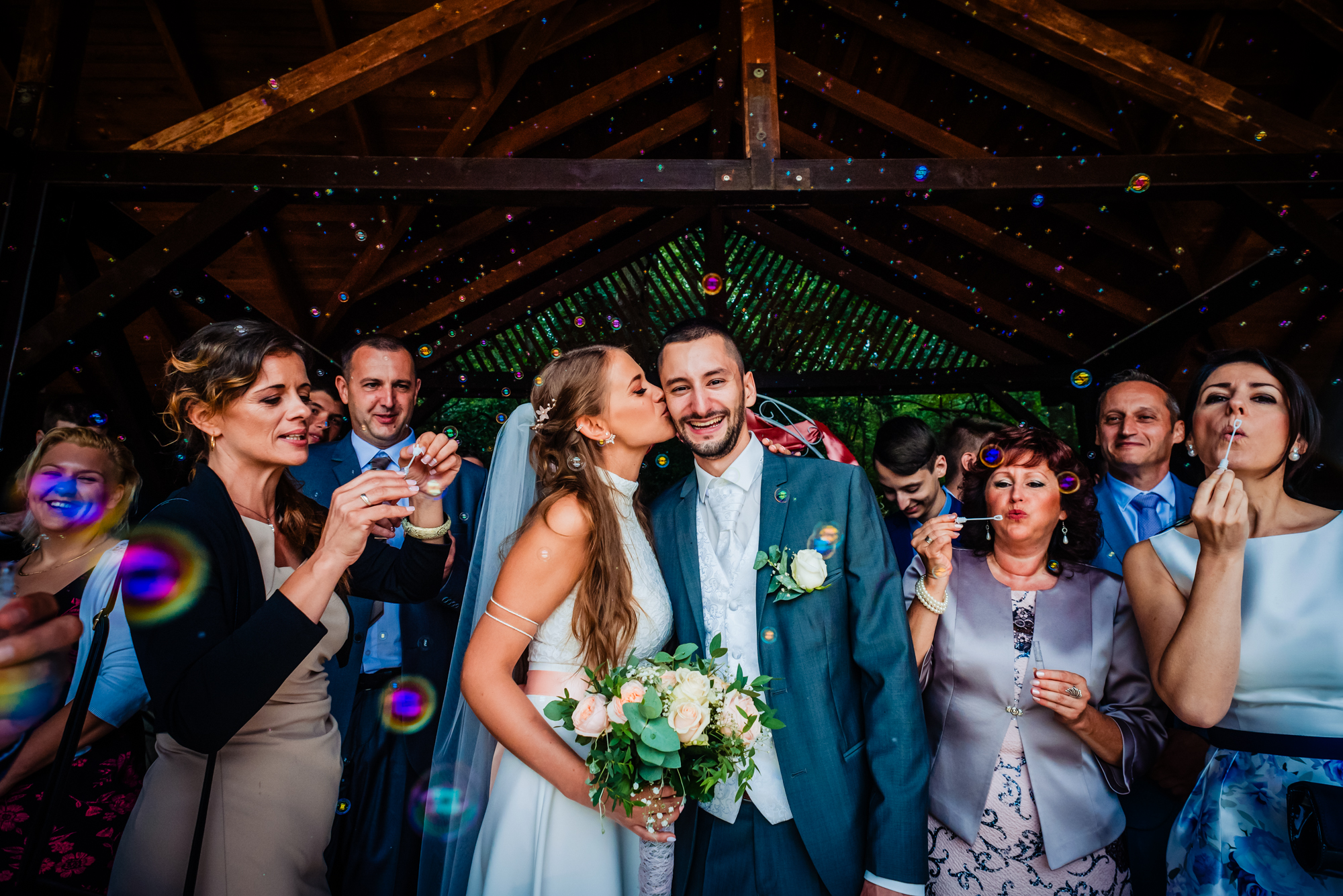 Bride kissing groom on the cheek as guests blow bubbles around them after the ceremony.