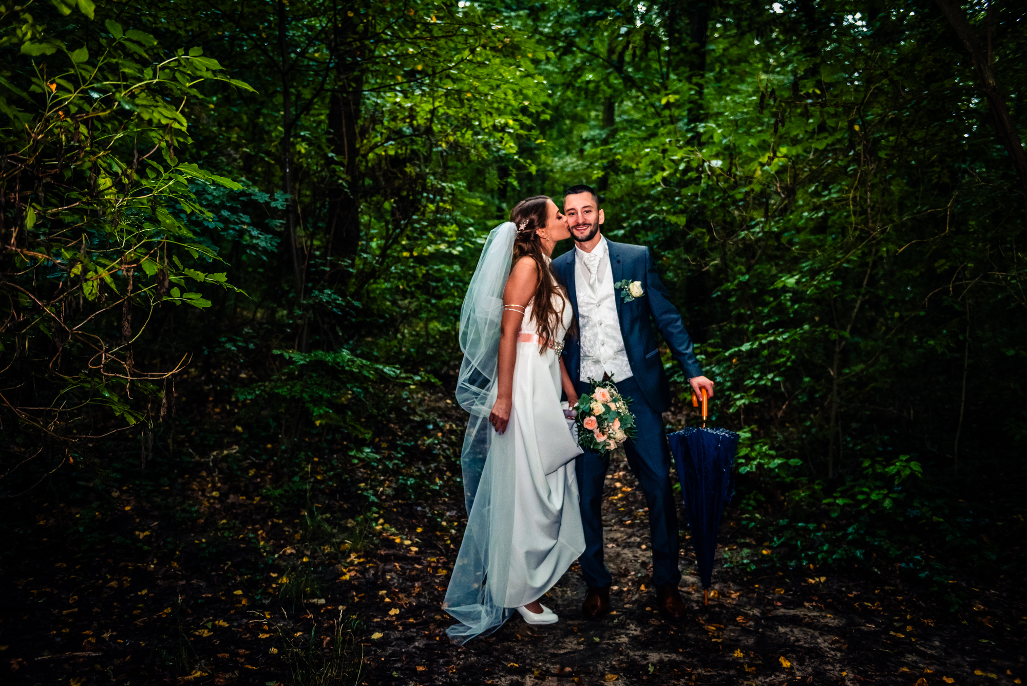 Bride kissing groom on muddy forest path while holding a closed umbrella after summer rain.