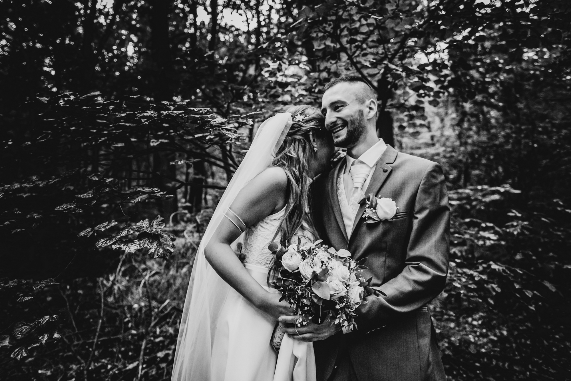 Bride hugging groom in forest while holding bouquet during black and white wedding portrait.