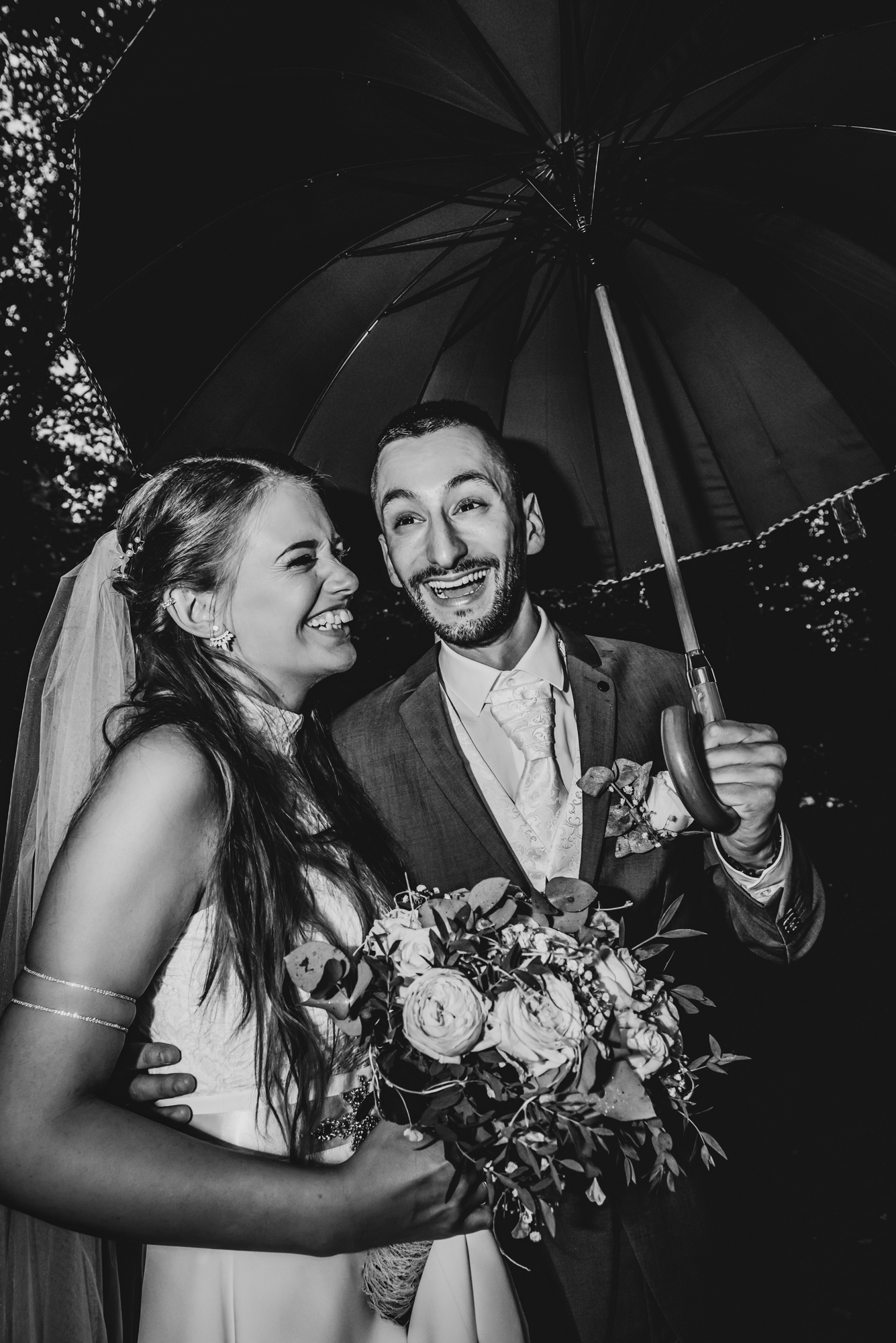 Bride and groom laughing together beneath umbrella during rainy outdoor wedding portraits.
