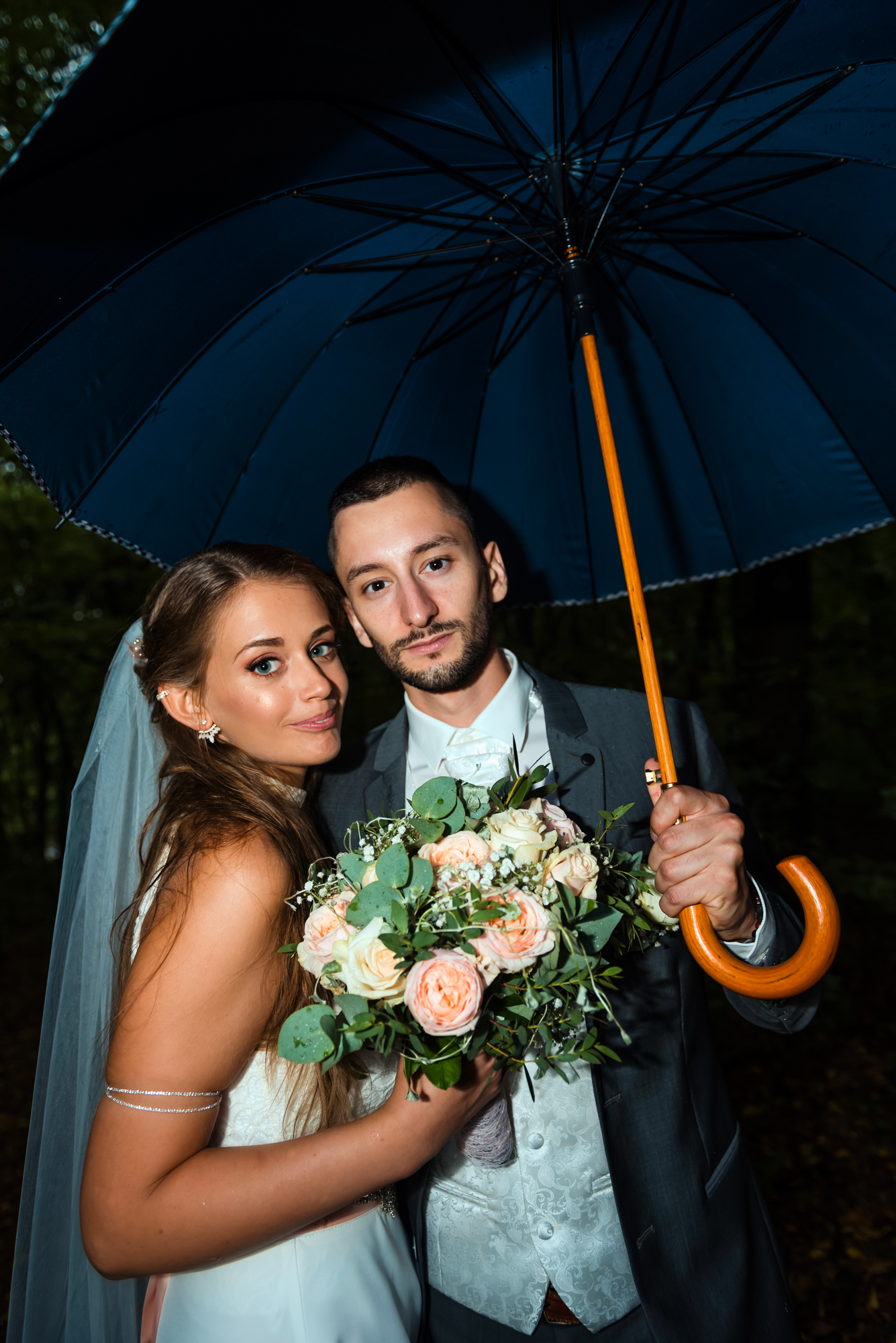 Bride and groom holding a bouquet beneath an umbrella during a rainy outdoor wedding portrait.