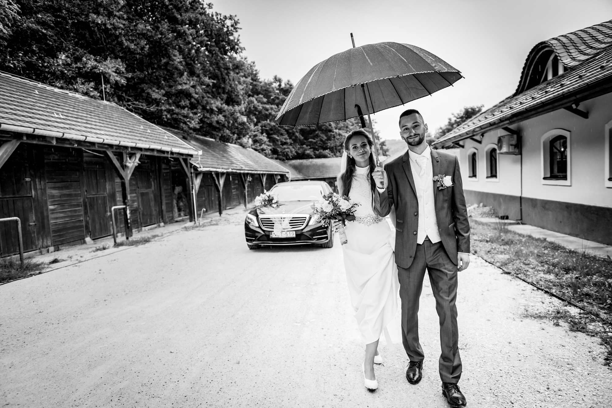 Bride and groom walking beneath umbrella beside wedding car on rainy countryside wedding day.