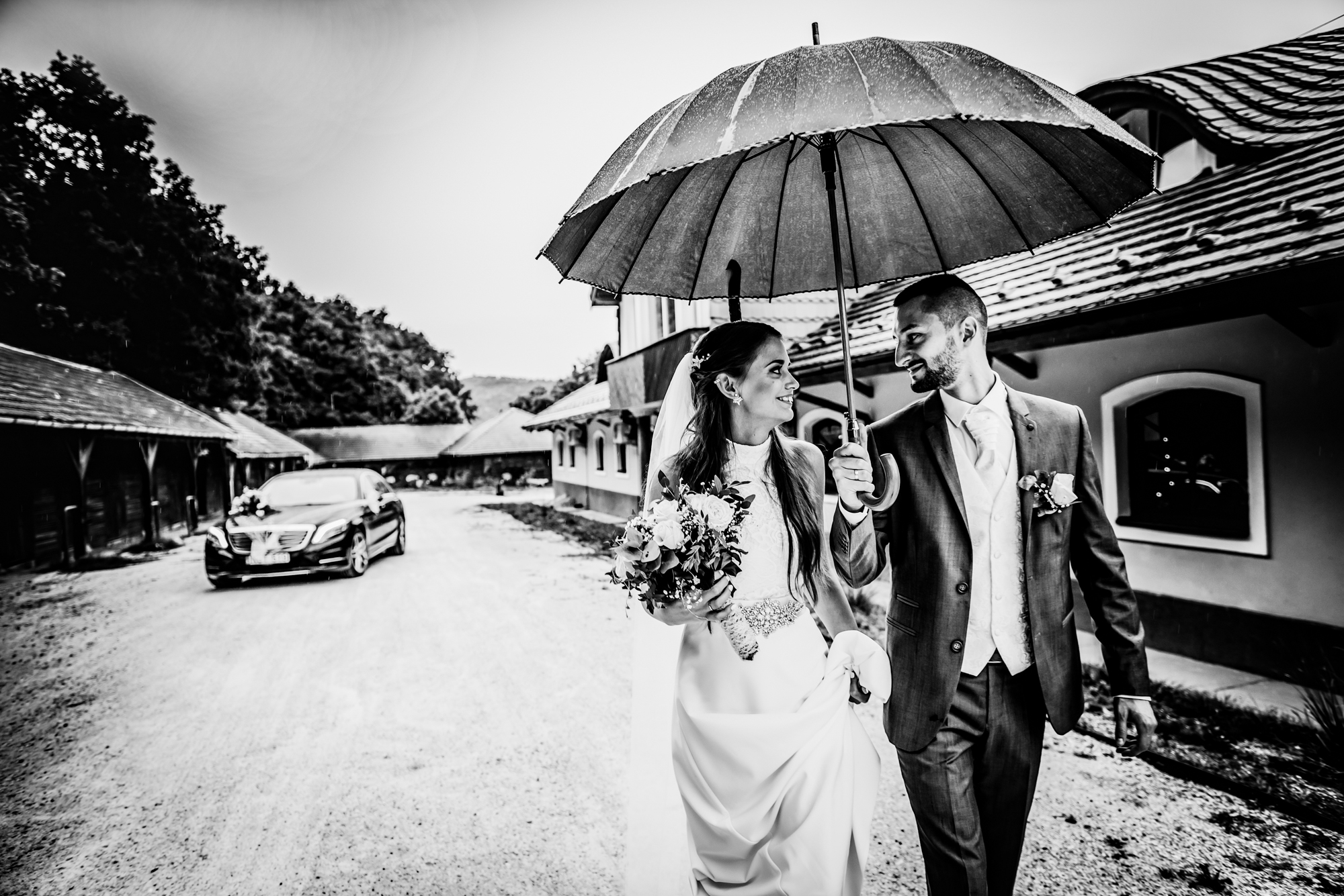 Bride and groom walking together beneath umbrella outside rustic countryside wedding venue.