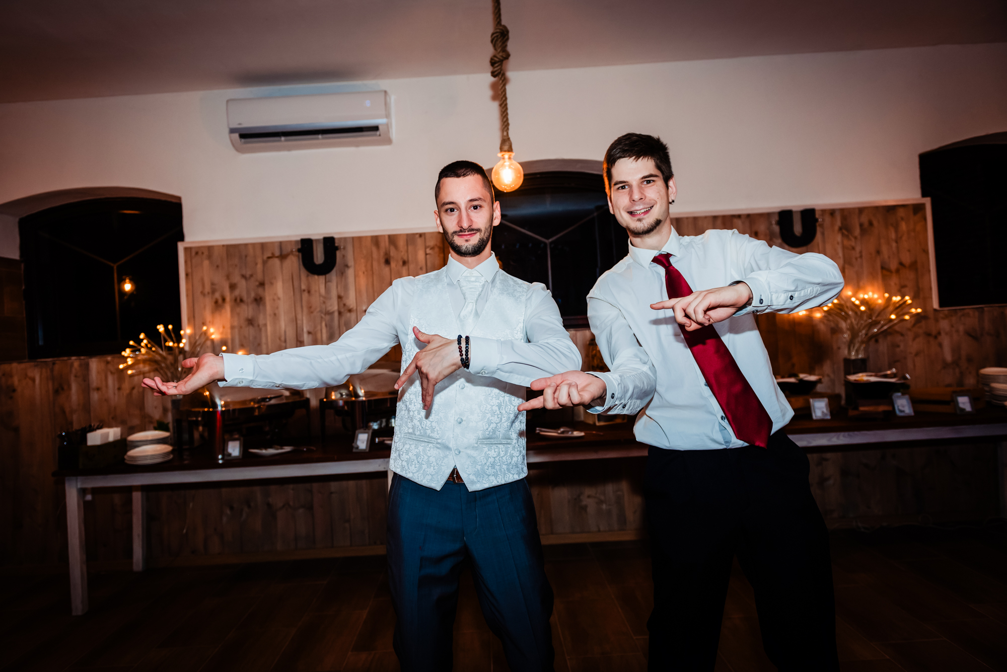 Groom and friend posing playfully together during evening wedding party at rustic indoor reception venue.