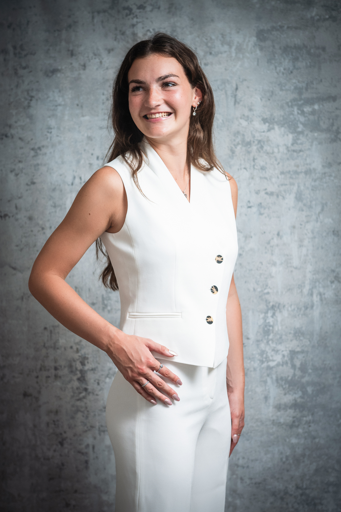Professional studio portrait of woman in white sleeveless blazer and wide-leg trousers against textured backdrop.