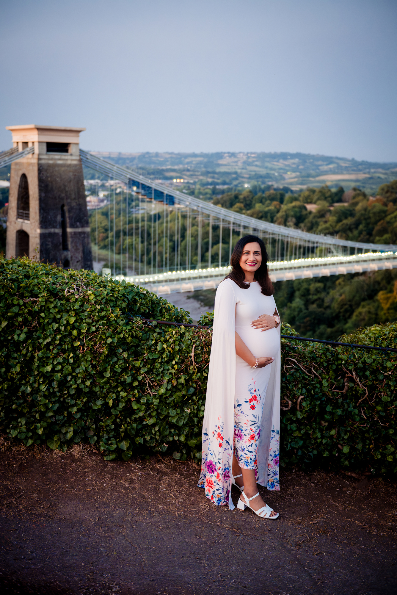Pregnant woman standing near Clifton Suspension Bridge viewpoint during sunset maternity photoshoot in Bristol.