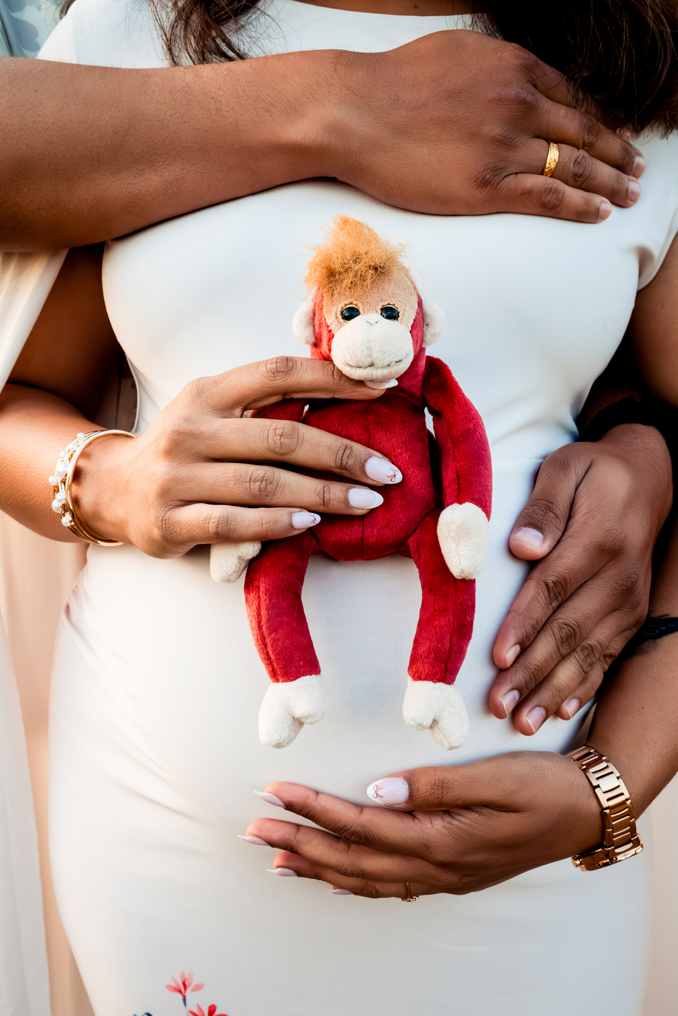 Hands holding baby bump with small red toy monkey placed on white maternity dress.