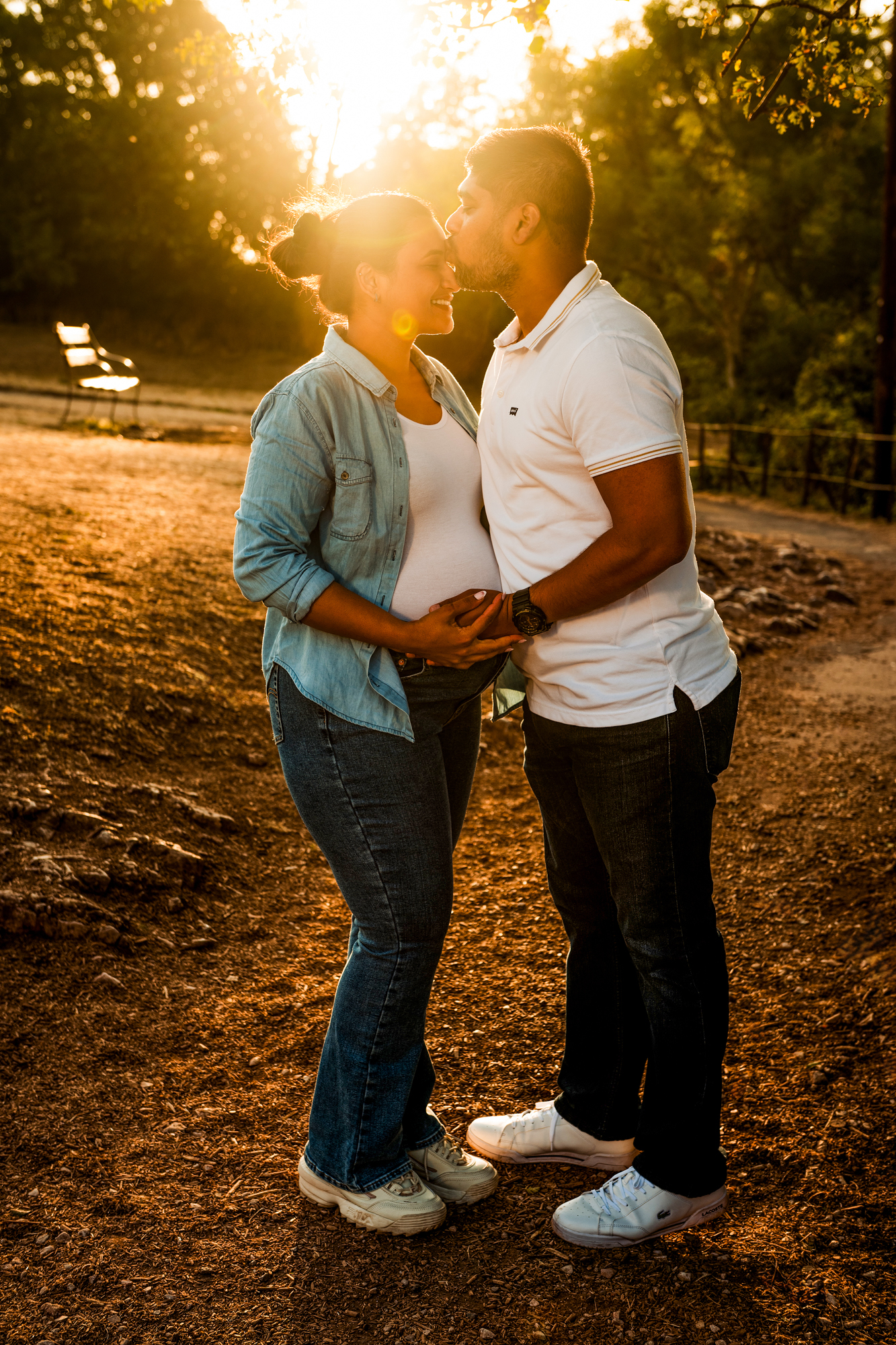 Pregnant couple sharing a kiss while holding baby bump in warm golden sunset light during outdoor maternity session in Bristol.