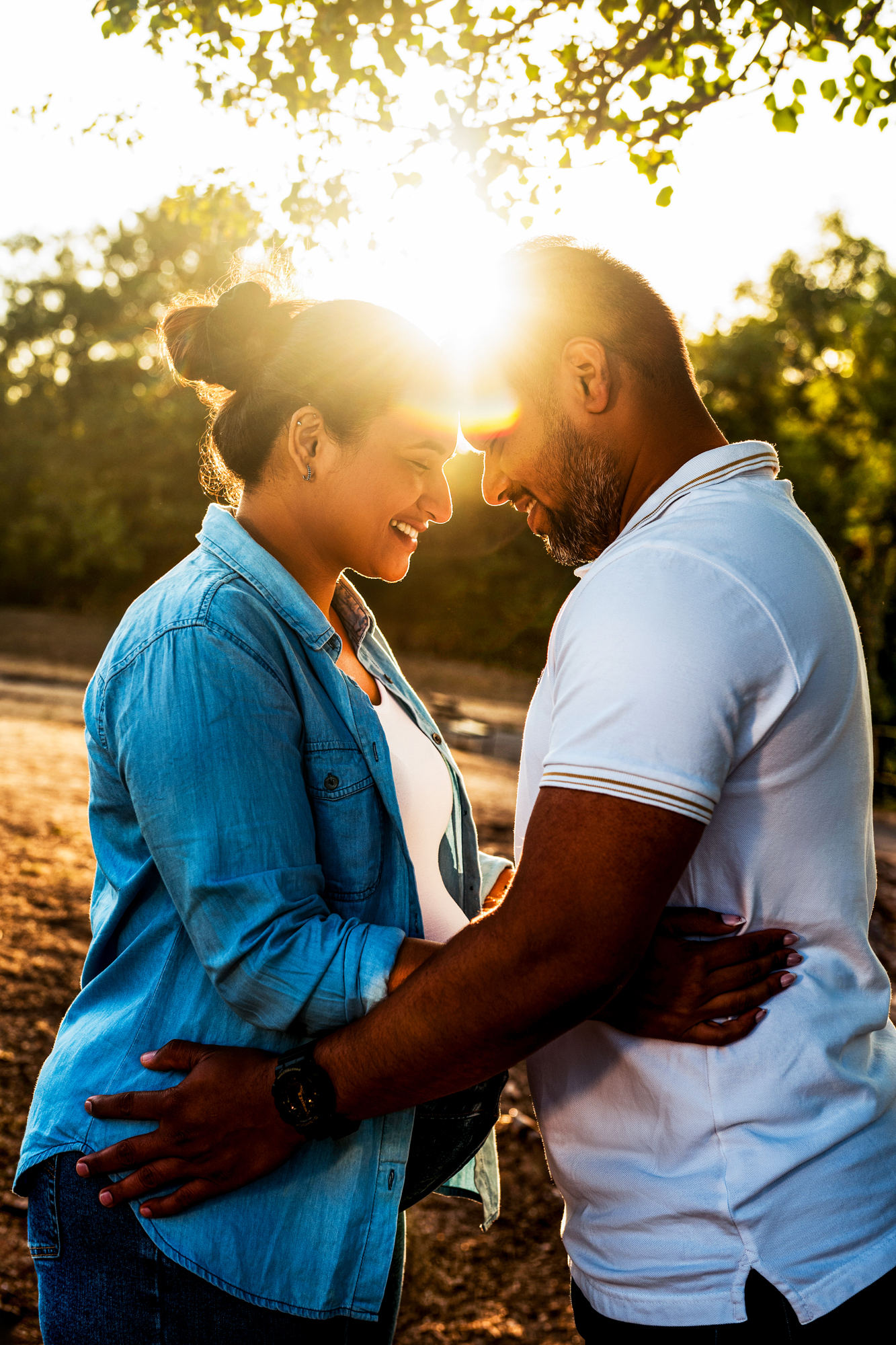 Close-up of couple touching foreheads with sunlight shining through trees during maternity session.