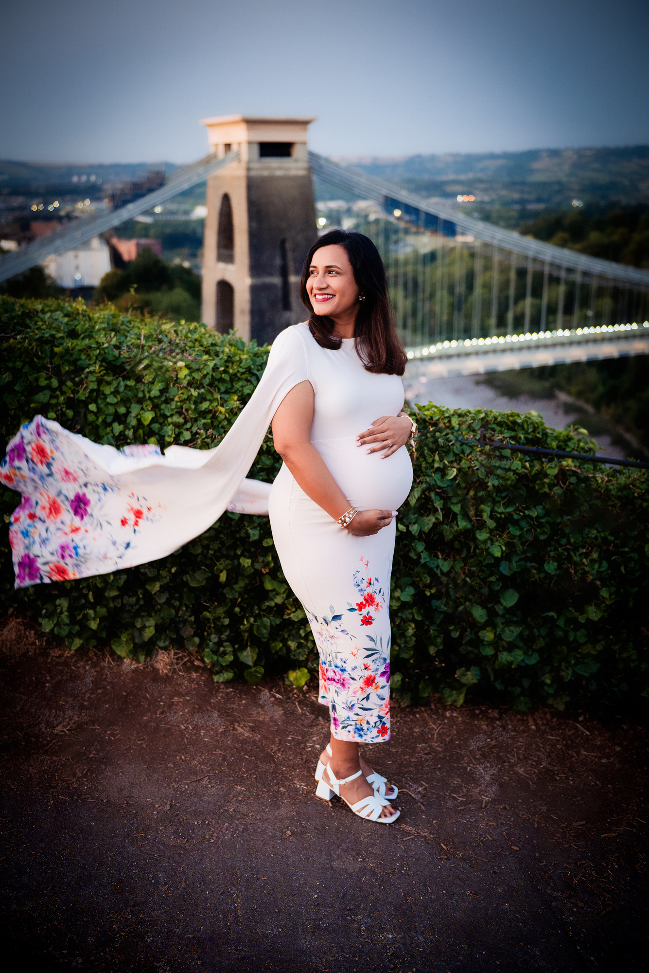 Pregnant woman in white floral dress overlooking Clifton Suspension Bridge at sunset in Bristol.