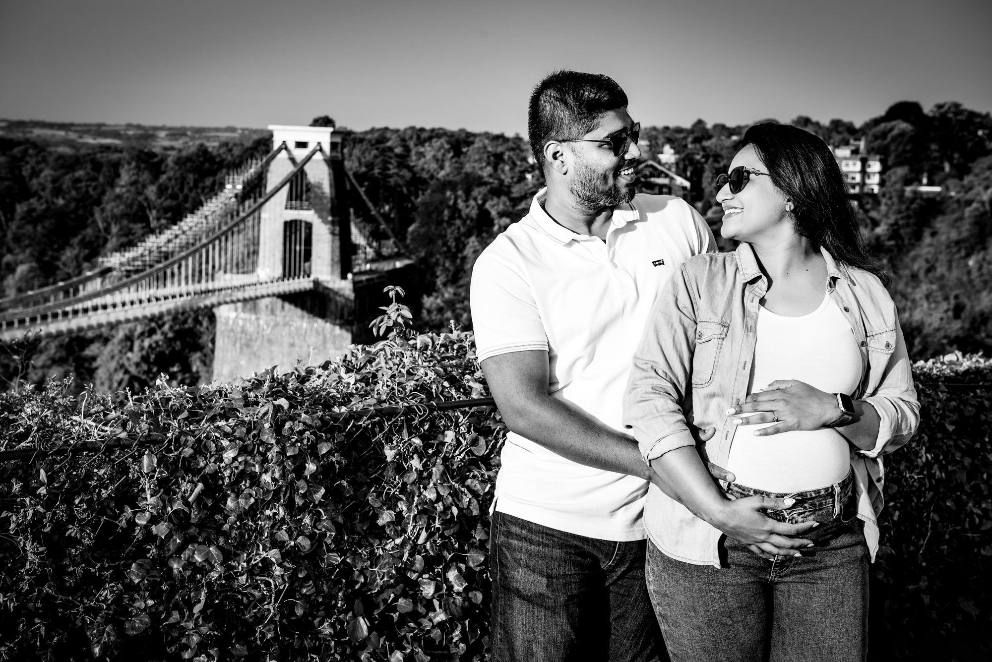 Couple smiling at each other while holding baby bump with Clifton Suspension Bridge behind them in black and white.