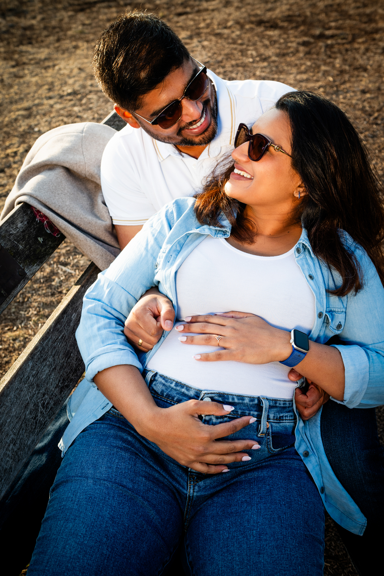 Expectant couple sitting on bench, smiling and holding baby bump during relaxed outdoor maternity session.