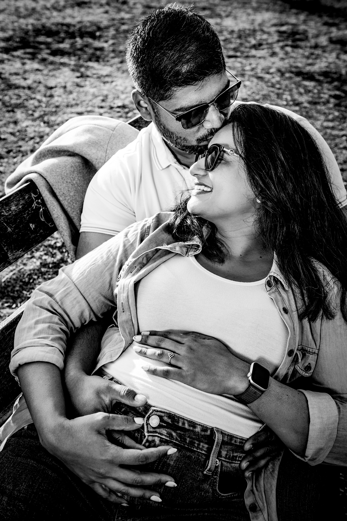 Black and white close-up of couple embracing on bench, hands resting on baby bump.