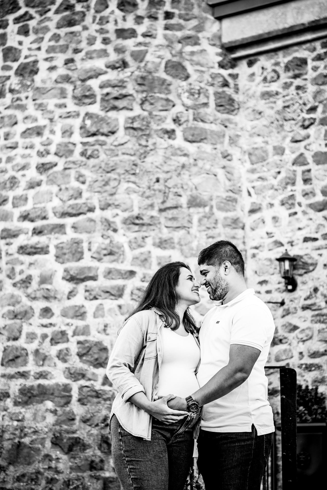 Couple standing close together against stone wall, smiling and holding baby bump during maternity shoot.