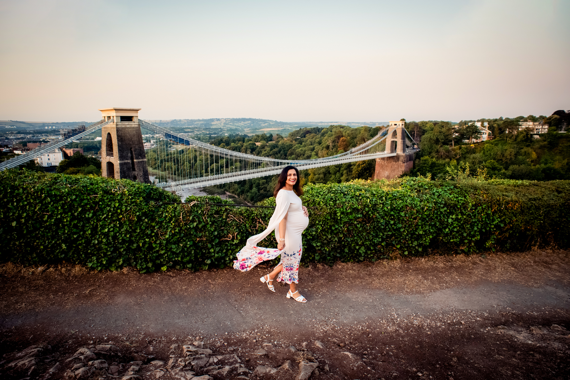Pregnant woman walking along path with Clifton Suspension Bridge in the background during golden hour.