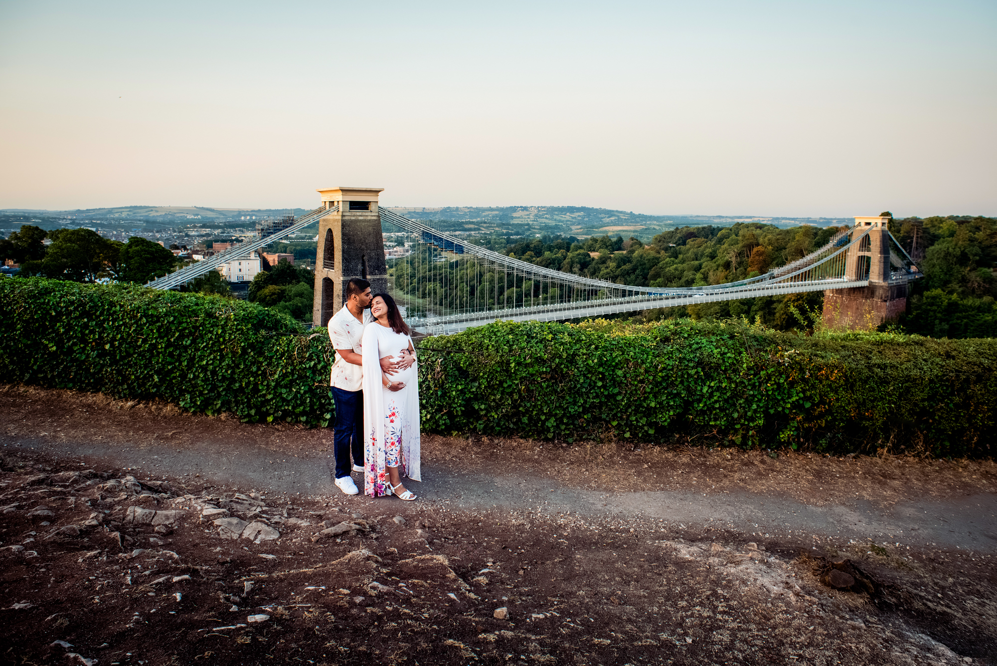 Wide landscape view of maternity session with Clifton Suspension Bridge and Bristol skyline behind couple.