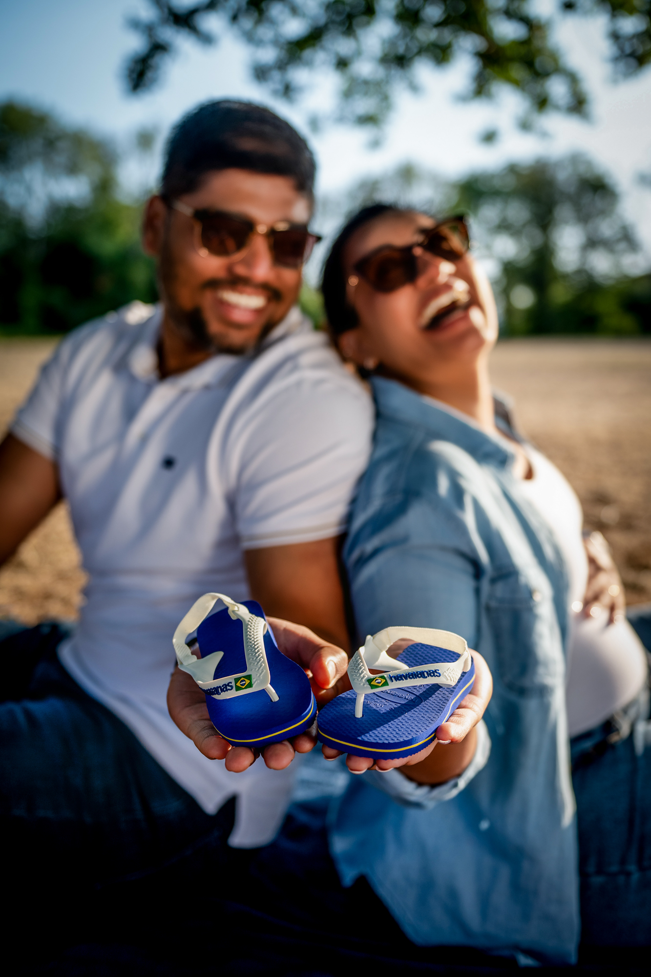 Couple holding tiny blue baby sandals toward camera during maternity session in warm evening light.