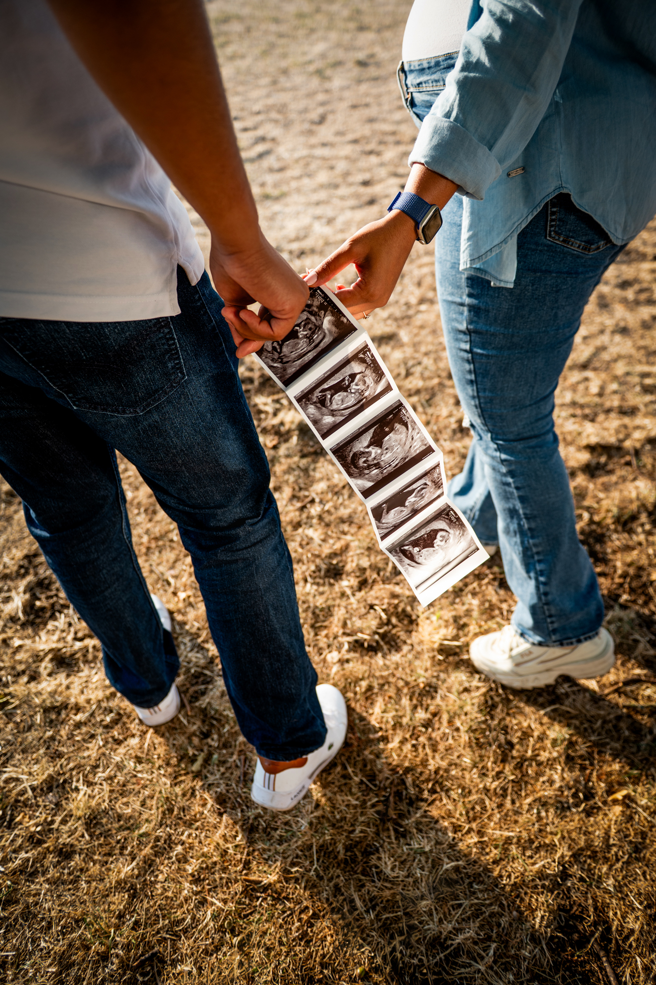 Parents holding ultrasound scan photos while walking together during outdoor maternity shoot.