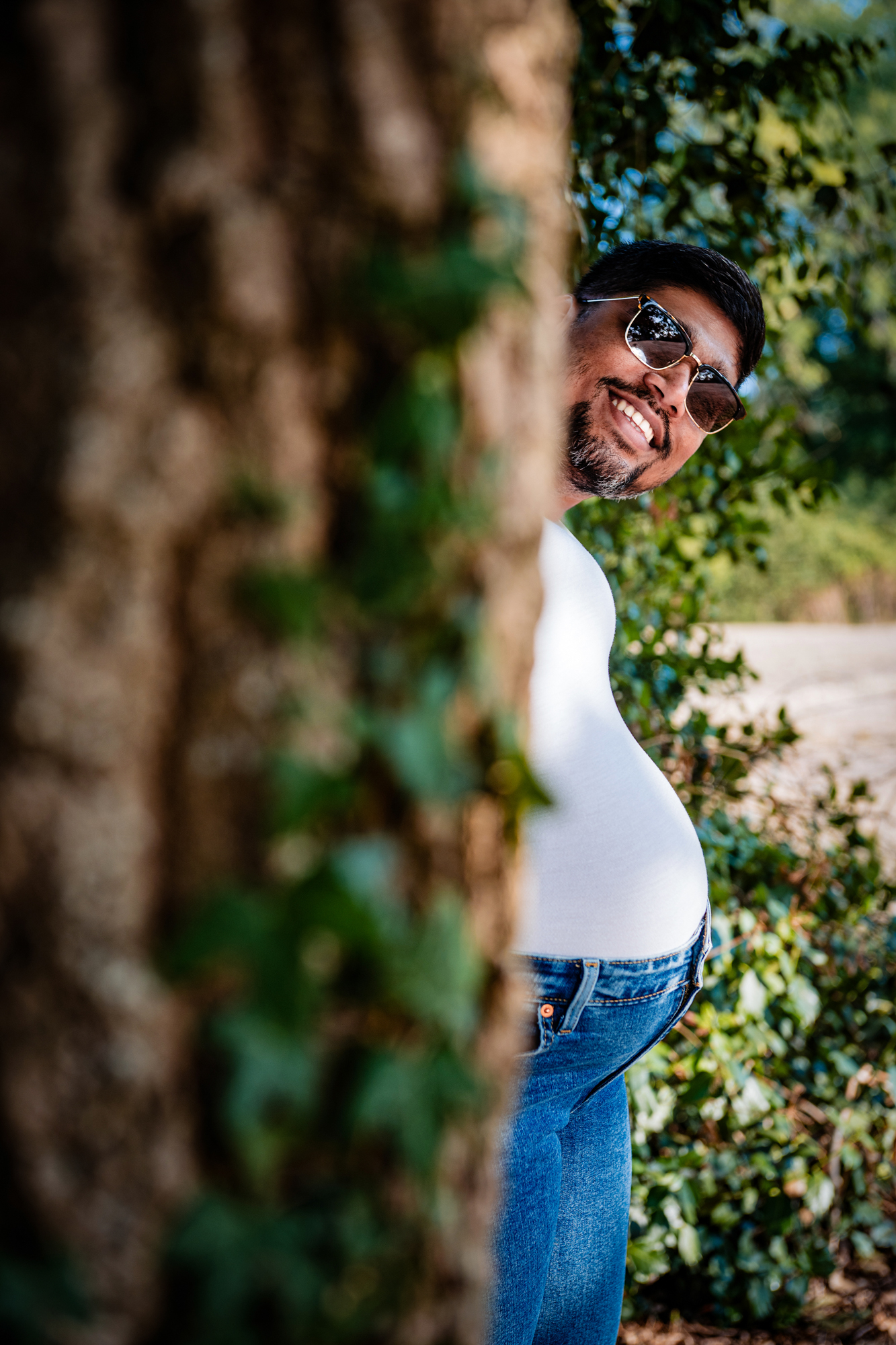 Partner smiling while peeking from behind tree during playful maternity photoshoot moment.