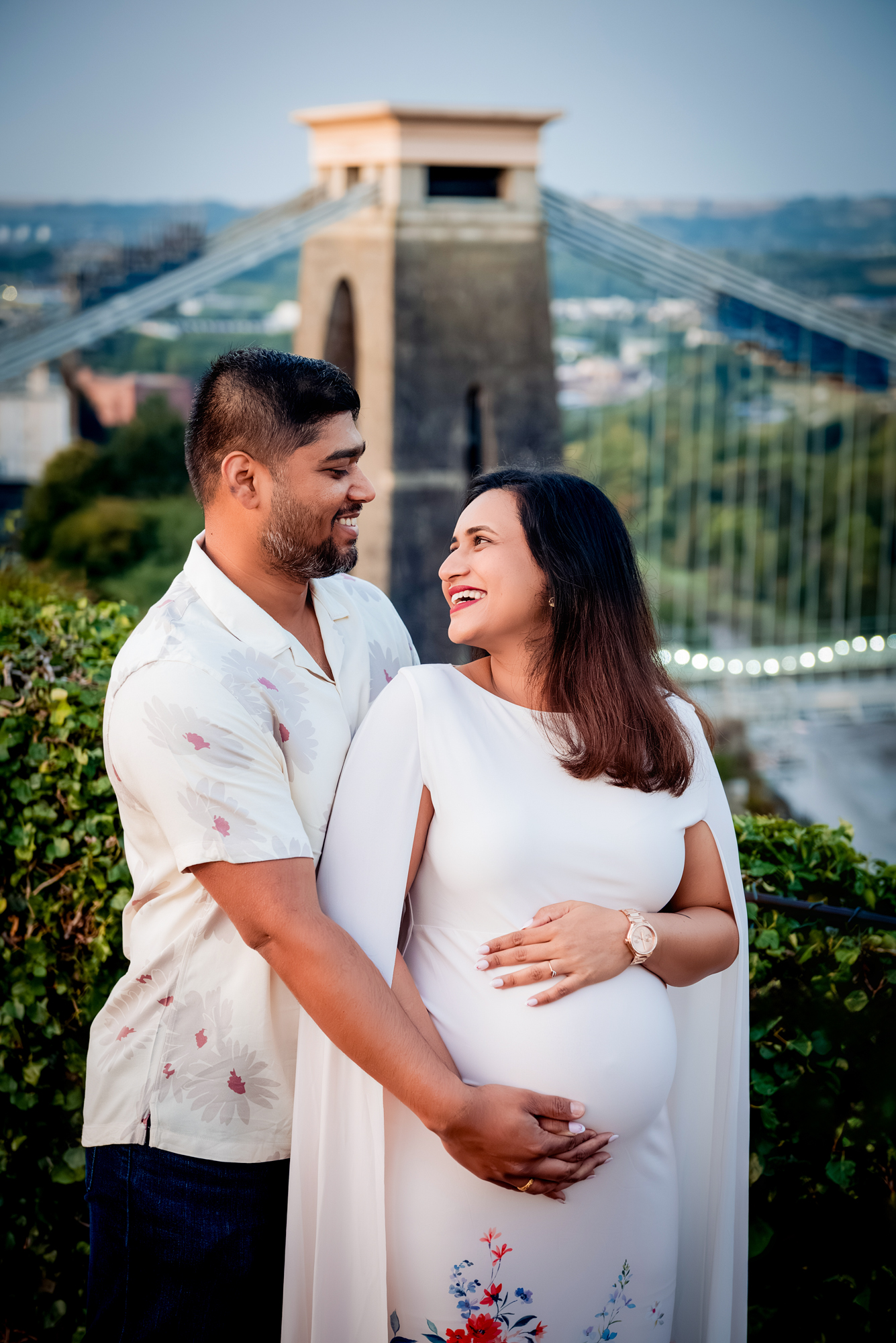 Couple smiling and holding baby bump with Clifton Suspension Bridge behind them at sunset.