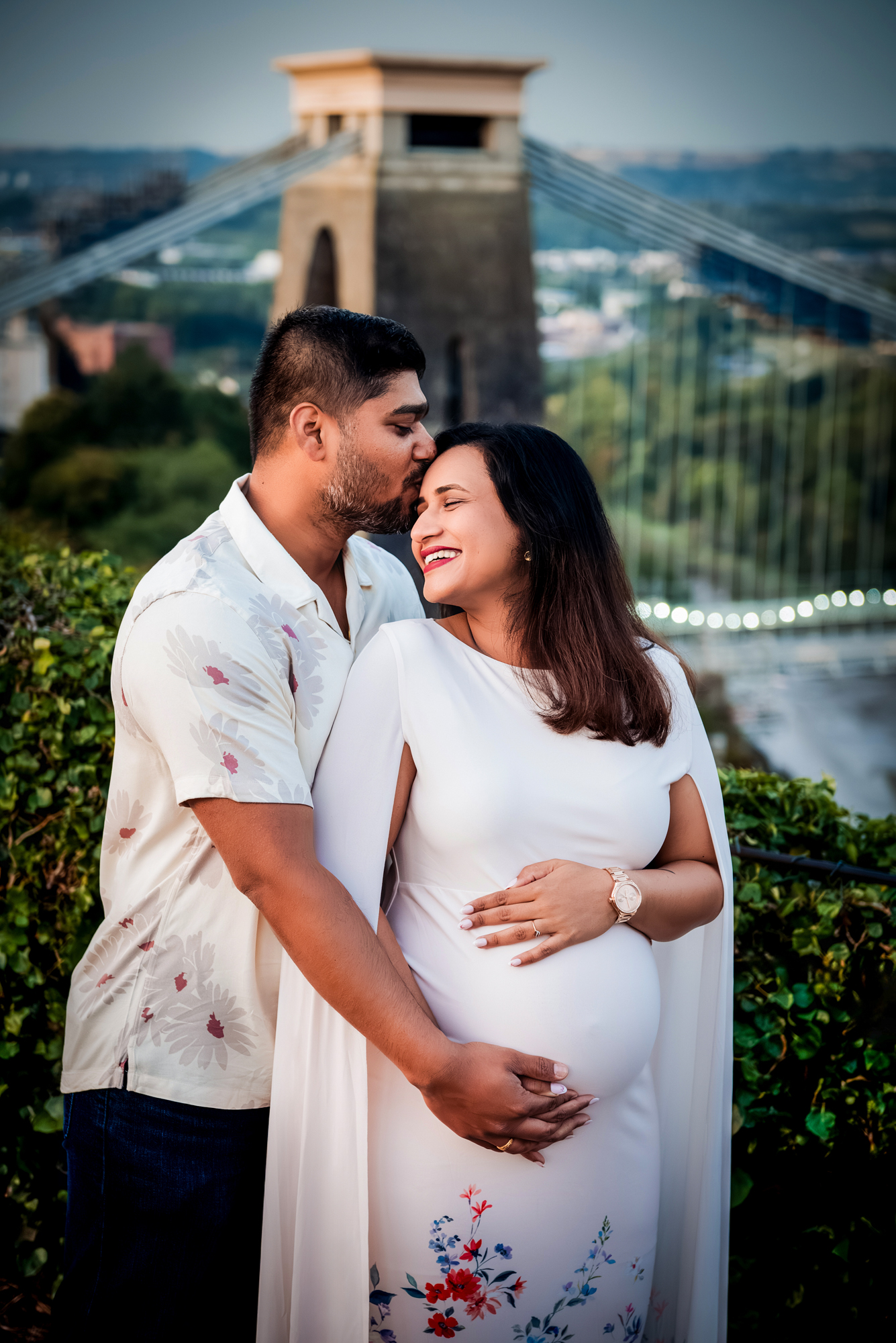Partner kissing pregnant woman’s forehead with Clifton Suspension Bridge in the background.