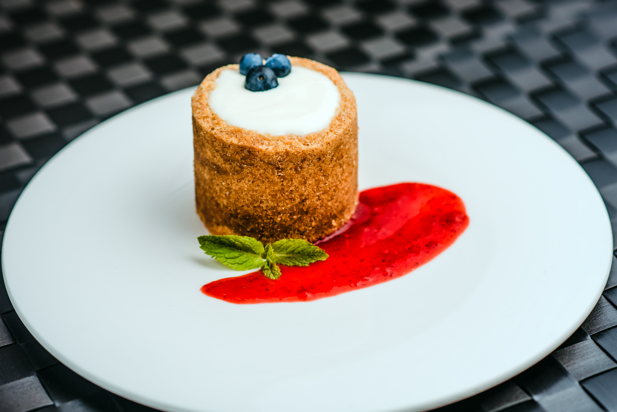 Mini cheesecake dessert with blueberry topping and berry coulis served on a white plate.