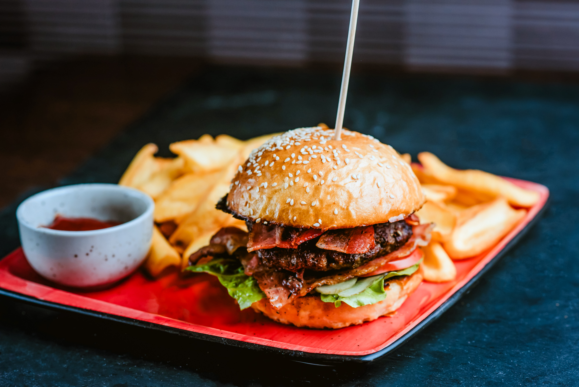 Bacon cheeseburger with fries and dipping sauce served on a red restaurant platter.