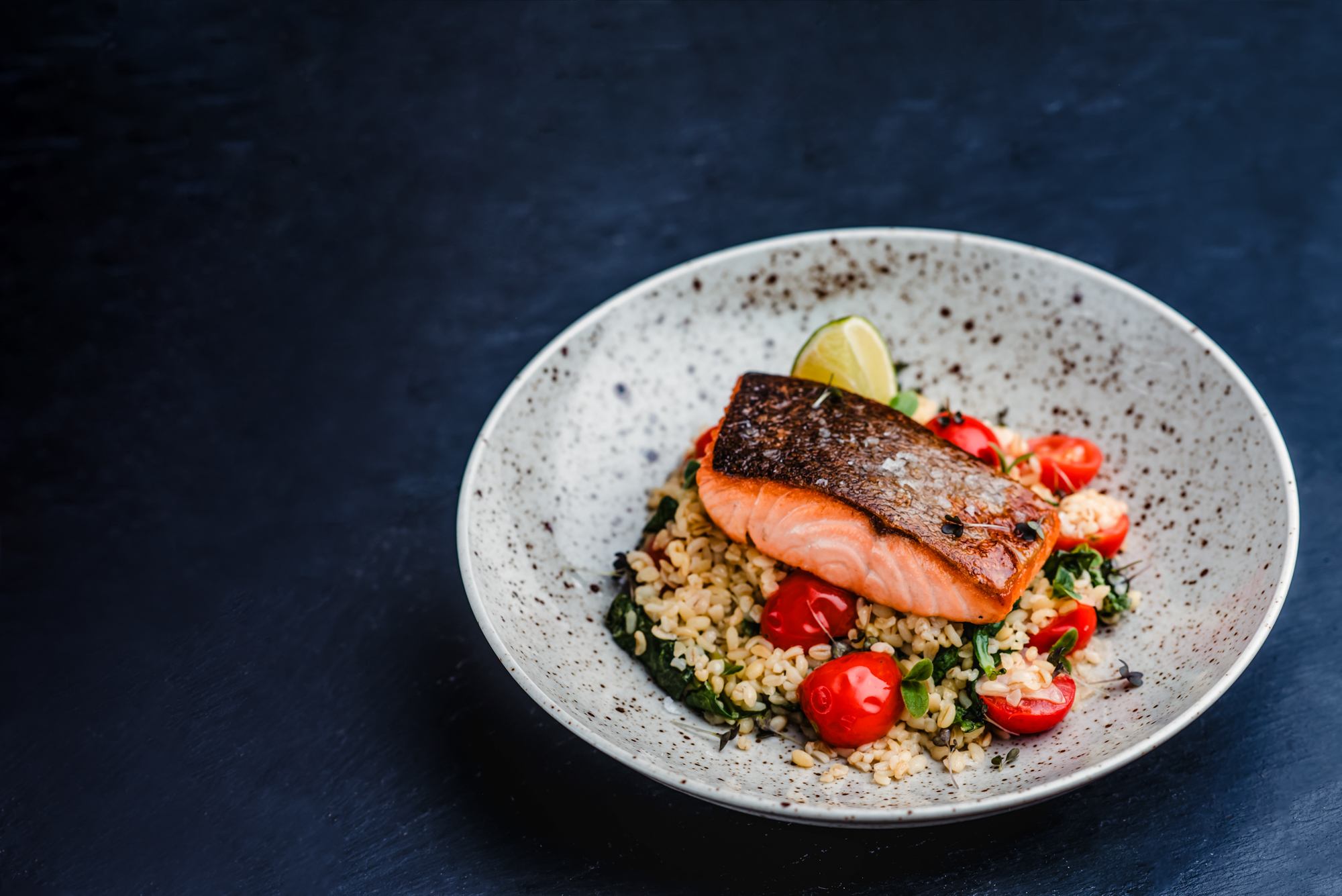 Crispy skin salmon fillet served with bulgur wheat, spinach, cherry tomatoes, and lime garnish.