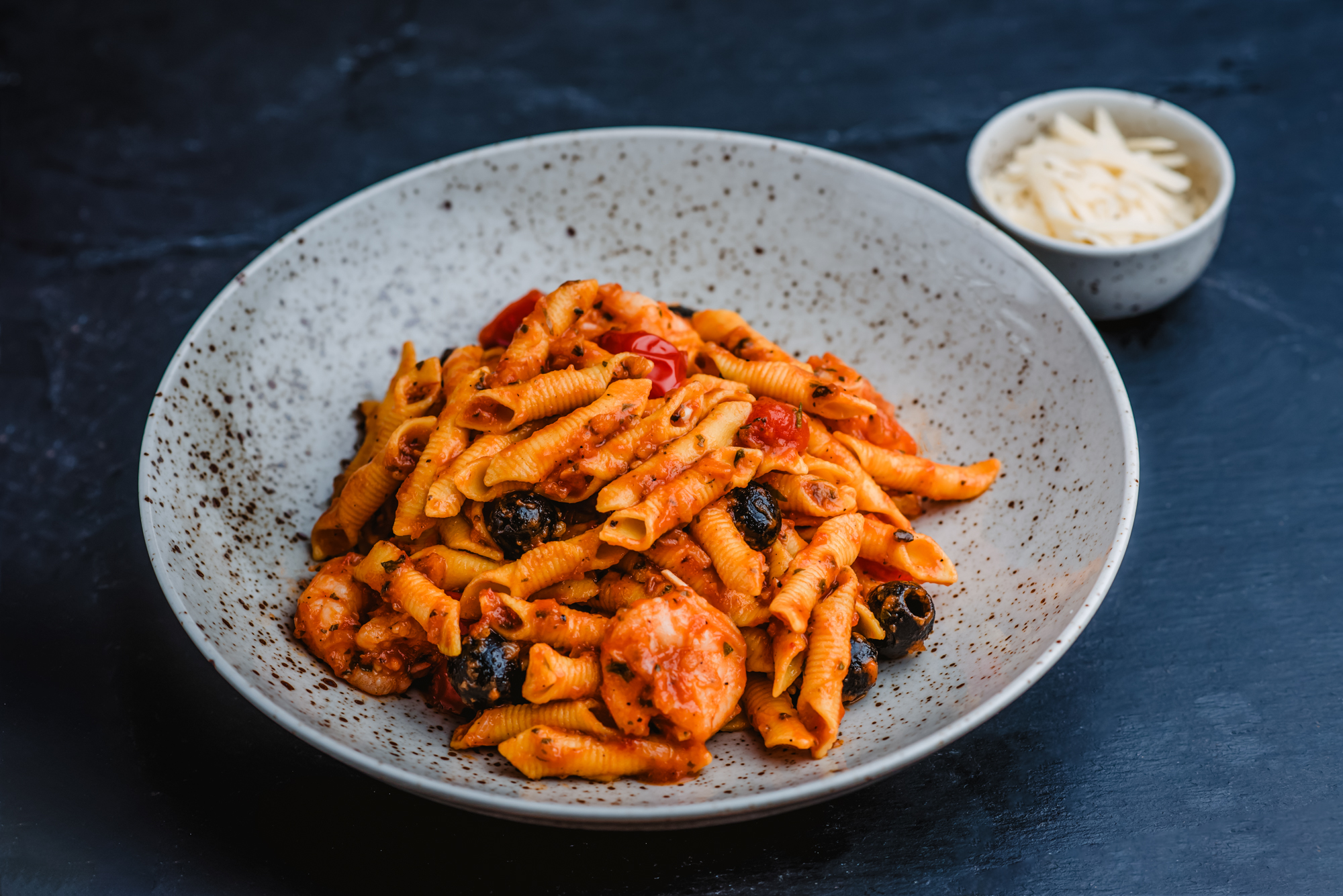 Seafood pasta with prawns, olives, tomato sauce, and grated parmesan served in a ceramic bowl.