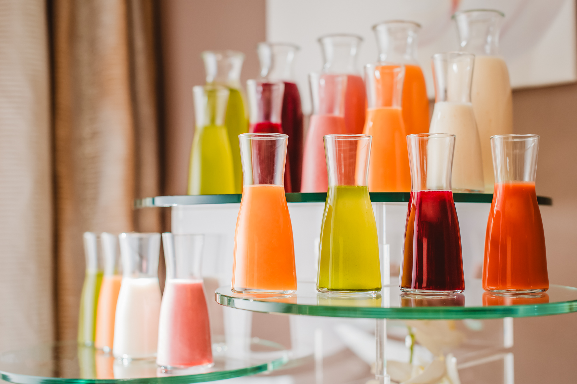 Assorted fresh fruit juices in glass bottles arranged on tiered glass stands at a hotel buffet.