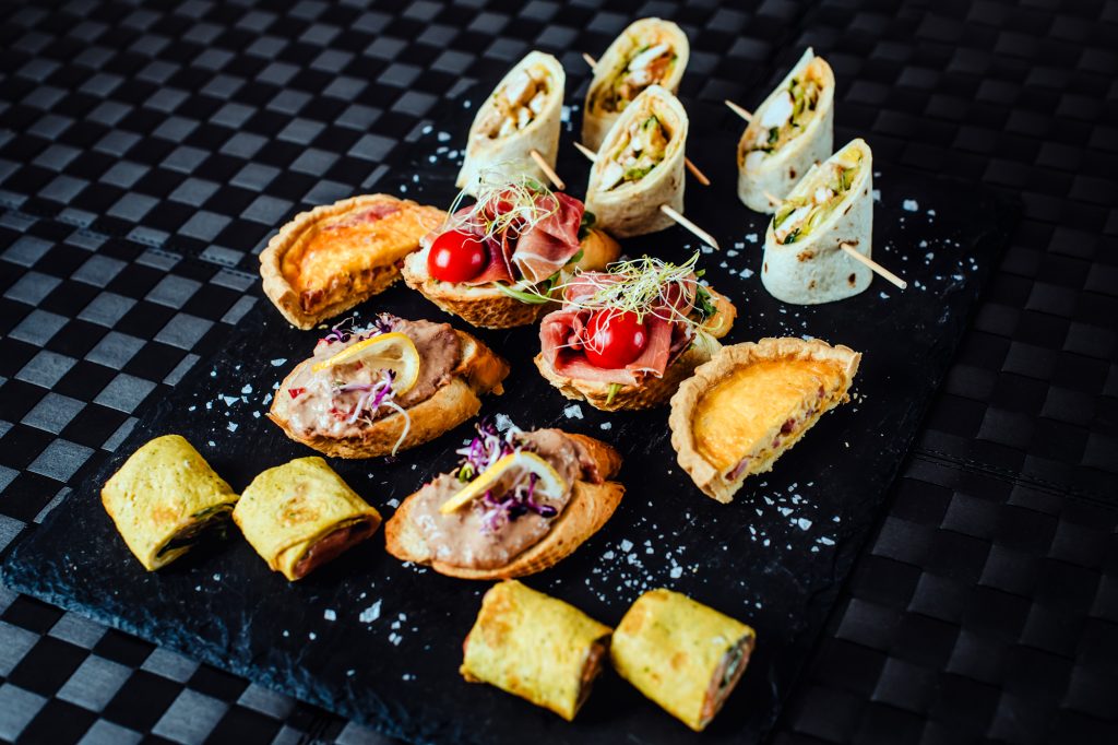 Assorted gourmet canapés and tortilla wraps displayed on a dark serving board for hotel catering.