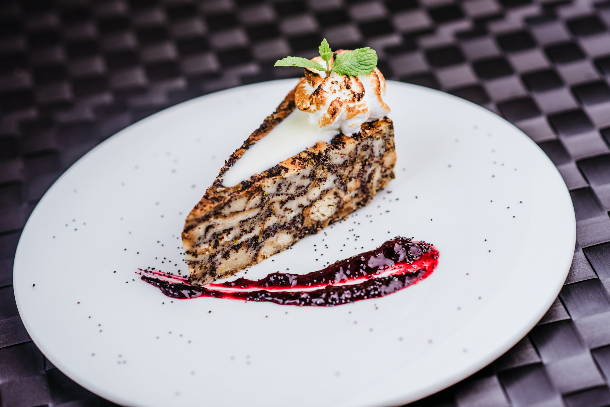 Slice of poppy seed cheesecake with berry sauce and toasted meringue garnish on a white plate.