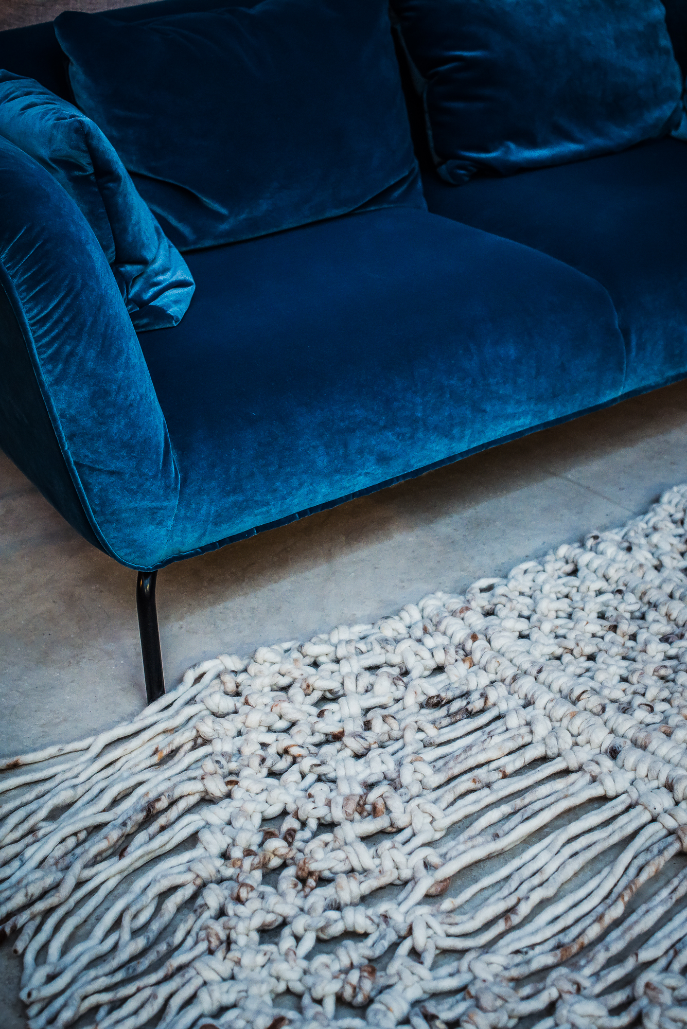 Textile product photography of blue velvet sofa beside thick woven tassel rug on polished concrete floor.